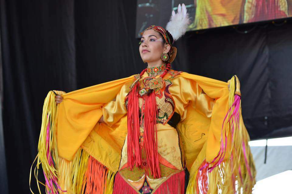 A young woman stands on a stage with her arms stretched out while wearing regalia